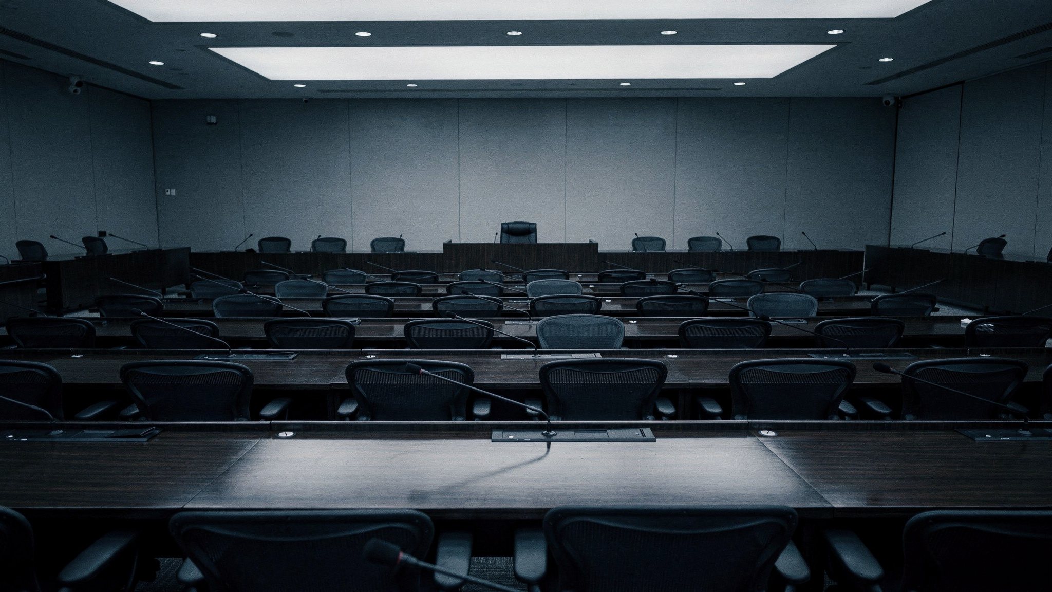 An empty government hearing room with rows of dark wooden desks and microphones under flat overhead light
